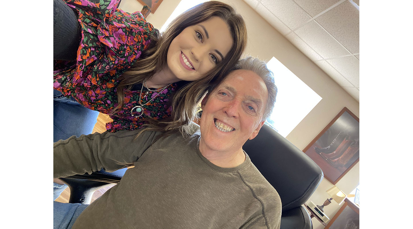 Shelbi Tidwell with brown hair, leaned over posing next to Rodney Ammons, an old man in a green shirt, smiling. 